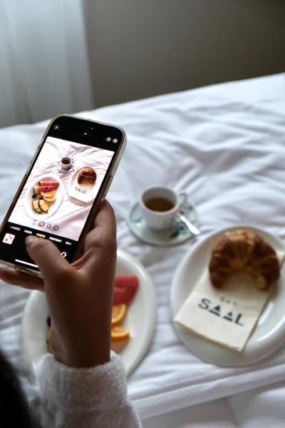 Gast fotografiert Fruehstueck im Bett mit Croissant und Kaffee