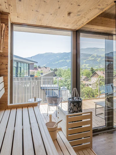 Finnische Sauna mit hellen Holzbänken, Aufgusseimer und Panoramafenster mit Blick auf die Berge und Außenterrasse